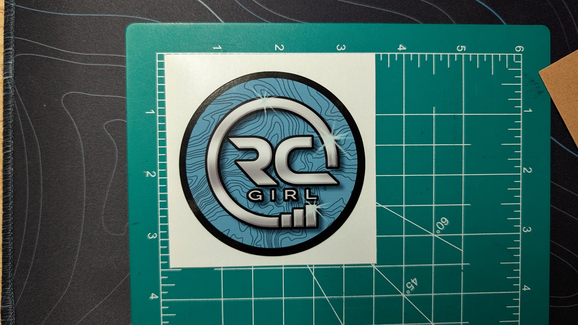 Circular logo with text on a white square, placed on a green cutting mat with measurements.
